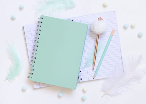 Light green spiral notebook on a white table near fur pen and pencil, feathers and pompoms, top view, Blank textbook mockup. Minimal school flat lay.