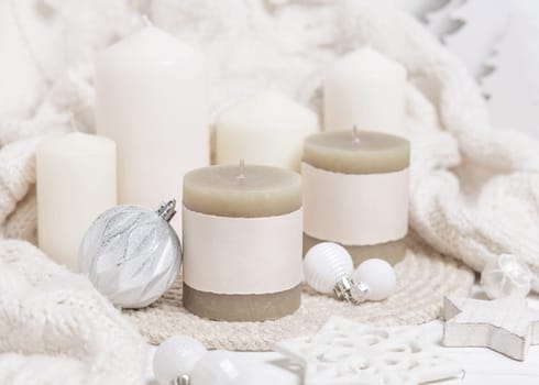 Pillar candles with blank label near white knitted sweater, wooden Christmas decor and lights, copy space. Winter atmospheric mock up in rustic or scandinavian style