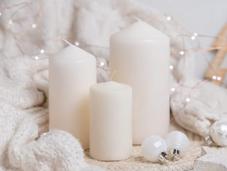 Pillar candles near white knitted sweater, wooden Christmas decor and lights, copy space. Winter atmospheric scene in rustic or scandinavian style