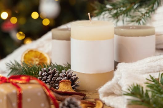 Pillar candles with blank label near Christmas decor, dryed fruits and fir twigs close up against Christmas tree lights, copy space. Warm winter family atmospheric mock up, magical Holiday time