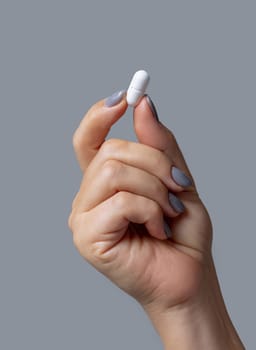 Female hand holding a white pill with fingers closeup against grey background. Healthcare, medical and pharmaceutical concept.