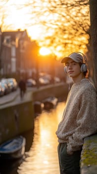 A young man leaning against a wall near the canals, wearing an oversized sweater and a baseball cap, listening to music through his vintage headphones. AI generative.