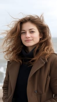 A woman with windblown hair stands by the sea, exuding confidence and elegance in a coastal setting.