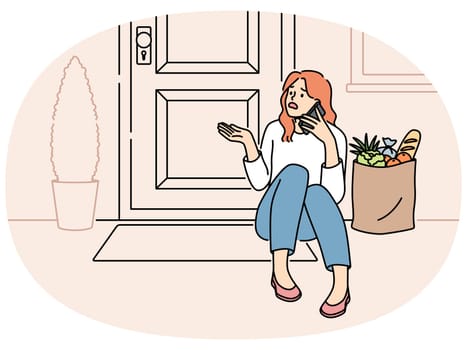 Embarrassed woman sits on threshold near door with jammed lock and cannot get into house, makes phone call. Puzzled girl with bag of groceries, calling specialist because of loss of key to door