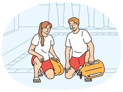 Pair of lifeguards sit by pool at hotel, holding swim training aids and keeping visitors safe. Smiling lifeguards or trainers working in fitness club urging people to visit swimming sports center