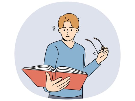 Man with book takes off glasses, trying to make out text in textbook and getting upset because of deterioration of vision. Guy is experiencing vision problems associated with excessive eye strain.