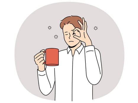 Sleepy man with mug of coffee, opens eyes with hand and needs to rest due to lack of energy and drowsiness. Tired young guy drinks coffee drink to cheer up and continue productive work.