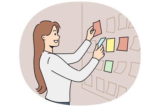 Business woman stands near scrum board with stickers and is engaged in kanban task planning. Girl manager or team leader of it company smiling while preparing new scrum sprint for programmers
