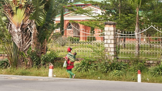 Moramanga, Madagascar - April 25, 2019: Unknown Malagasy woman carrying her baby on back in wrap scarf, luxury house in background. Most people at Madagascar are poor, and only very few wealthyMadagascar - April 25, 2019:
