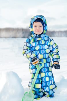 Child having fun playing with snow on sunny winter day, wearing colorful snowsuit and holding a green shovel