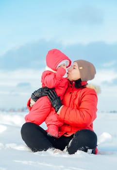 Mother is kissing her baby while sitting on snow during beautiful winter day