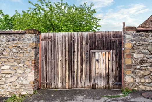 A rustic wooden gate stands tall between aged stone walls, with a smaller door adjacent. Lush green trees frame the entrance under a clear blue sky, creating a serene atmosphere4