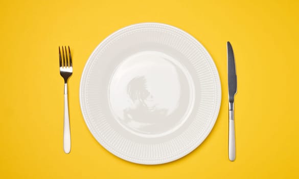 Round white empty ceramic plate and knife with fork on yellow background, table setting. Top view