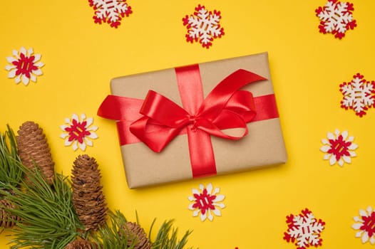 Gift box with red satin ribbon and bow on a yellow background. Holiday background. There is a pine branch with cones nearby