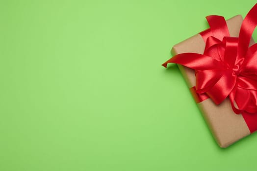 Box wrapped with red ribbon and brown craft paper on a green background, top view. Copy space