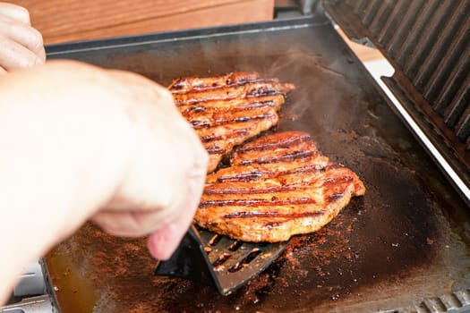 Two pork cutlet steak grilled on electric grill, blurred woman hand holding plastic spatula, about to turn one piece of meat
