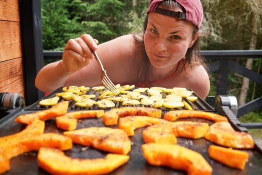 Young woman moving pieces of potato with fork, blurred pieces of orange butternut squash grilled on electric grill in foreground
