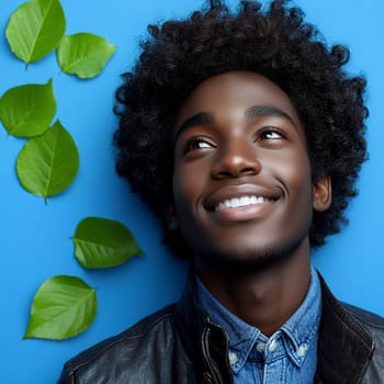 Stylish young man in leather jacket and denim shirt against blue background with green leaves, natural cheerful portrait with bright smile.