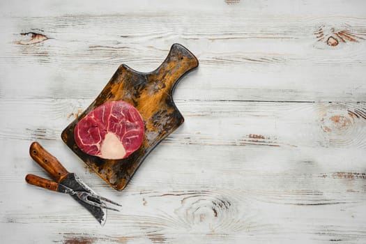 A raw beef steak on a wooden cutting board. Butcher knife and fork are placed below the cutting board on a white wooden tabletop