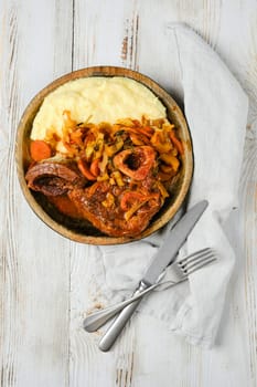 A wooden bowl filled with osso buco, mashed potatoes, and vegetables. A knife and fork are placed on a white napkin to the right of the bowl.