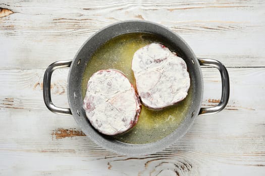 Overhead view of two floured steaks are sizzling in a pan with oil. The pan is on a white wooden tabletop