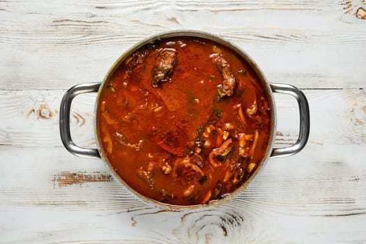 Overhead view of a silver pot with two handles on a white wooden table. The pot is filled with a rich beef stew, filled with chunks of meat and vegetables.