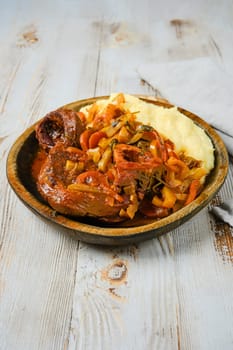 Osso buco, a traditional Italian dish, is served on a wooden plate with mashed potatoes, onions, peppers, and a rich gravy.