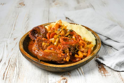 A wooden plate filled with osso buco, mashed potatoes, and vegetables. The osso buco is in a red sauce, and the mashed potatoes are on the side.
