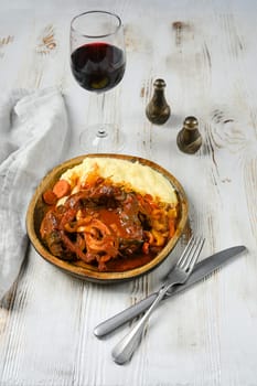 A wooden bowl with a red-brown beef stew, garnished with sliced onions and carrots, sits next to a mound of mashed potatoes. A glass of red wine and silverware are also on the white, wooden table.