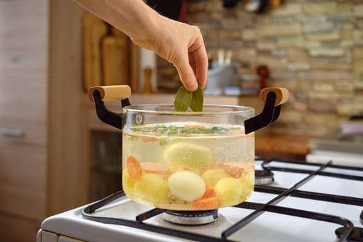 A hand is adding fresh bay leaves to a pot of simmering water with various vegetables, including potatoes and carrots, on a stovetop. The kitchen has a cozy, rustic ambiance.