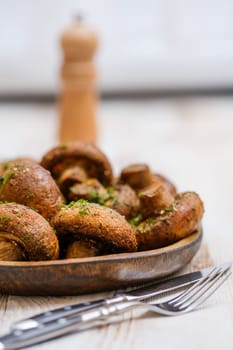 A wooden plate displays a heap of freshly cooked mushrooms seasoned with herbs, accompanied by a fork and knife. A pepper grinder sits in the background, suggesting a rustic dining setting.