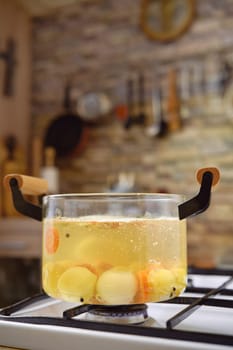 A clear pot filled with boiling water, potatoes, and carrots sits on a stovetop.