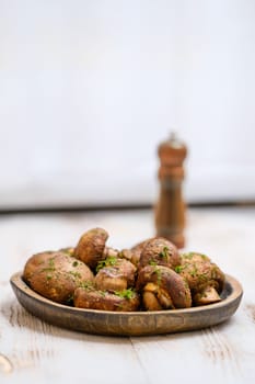 A wooden plate filled with roasted mushrooms garnished with herbs on a rustic table. A pepper mill in the background adds a touch of charm to this appetizing arrangement.