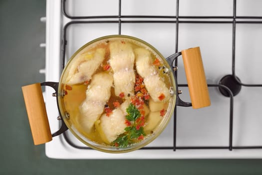 A pot of white fish fillets simmers slowly in clear broth, garnished with herbs and spices. The scene captures a warm, inviting kitchen atmosphere, emphasizing the home-cooked meal preparation.