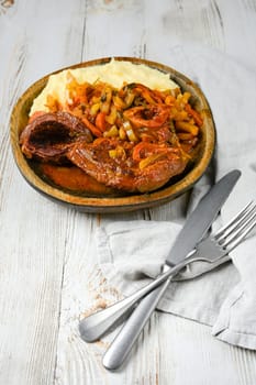 A wooden plate of osso buco served with mashed potatoes, cooked in a tomato and onion sauce with herbs. A silver knife and fork are placed beside a linen napkin on a white wooden table.