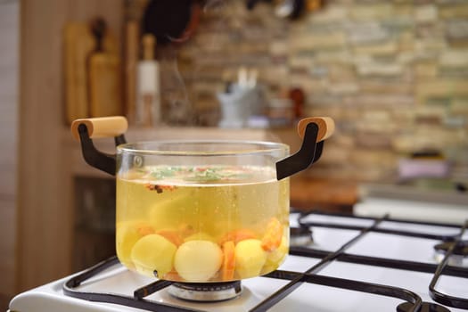 A clear pot filled with simmering vegetable broth sits on a gas stove, showcasing vibrant vegetables and herbs. The cozy kitchen features a rustic backdrop with wooden utensils and soft light.