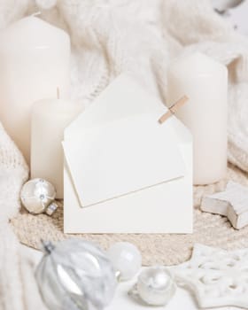 Blank paper card and envelope near white knitted sweater, pillar candles and Christmas baubles and decor, copy space. Winter atmospheric mock up for family greeting