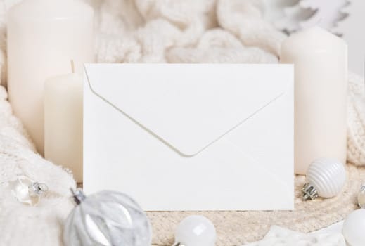 White blank envelope near white knitted sweater, candles and Christmas decor, copy space. Winter atmospheric mockup, family greeting in rustic or scandinavian style