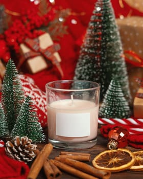 Glass candle with blank label near red knitted sweater and Christmas decor against lights and wrapped presents, copy space. Warm winter family atmospheric mock up, Holiday time
