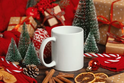 White ceramic coffee mug near Christmas decor, presents, trees, spices and red knitted sweater on wooden table closeup, copy space. Winter atmospheric mock up, holiday time