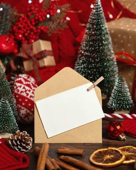 White postcard and craft envelope near Christmas decor, trees, spices and red knitted sweater on wooden table closeup, copy space. Winter atmospheric mock up, holiday greeting