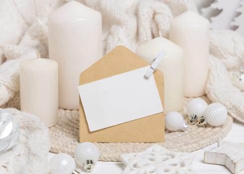 Blank paper card and envelope near white knitted sweater, pillar candles and Christmas baubles and decor, copy space. Winter atmospheric mock up for family greeting
