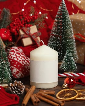Pillar candle with blank label near red knitted sweater and Christmas decor against lights and wrapped presents, copy space. Warm winter family atmospheric mock up, Holiday time