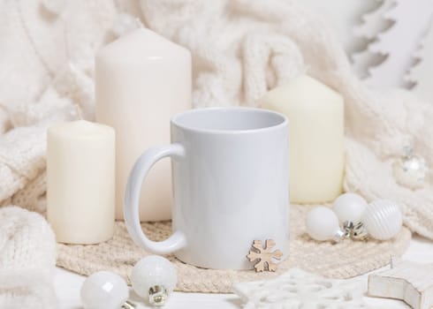 White blank coffee mug near candles, white knitted sweater and Christmas decor, copy space. Winter atmospheric mockup in rustic or scandinavian style