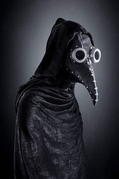 Plague doctor in hooded cloak at night over dark misty background