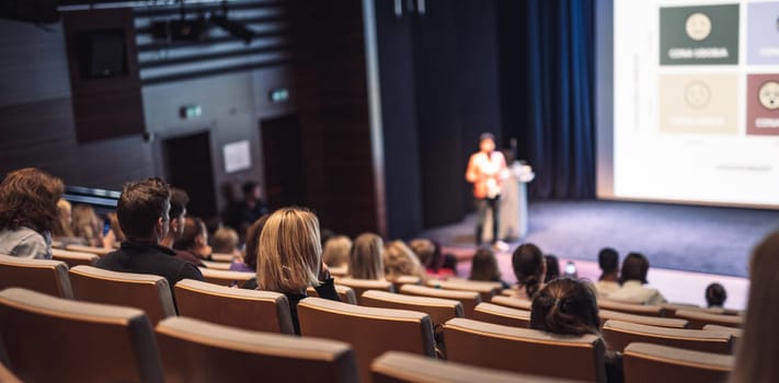 Business and entrepreneurship symposium. Female speaker giving a talk at business meeting. Audience in conference hall. Rear view of unrecognized participant in audience.