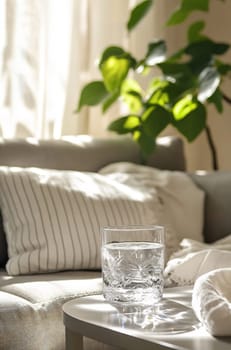 A clear glass of water rests on a light table beside a comfortable couch decorated with soft pillows, illuminated by natural light.