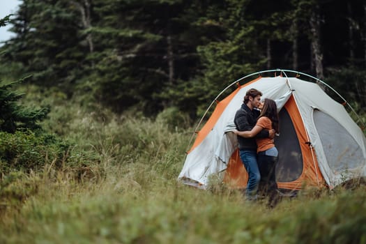 A couple shares an intimate moment outside their camping tent in a lush forest, surrounded by tall trees and greenery.
