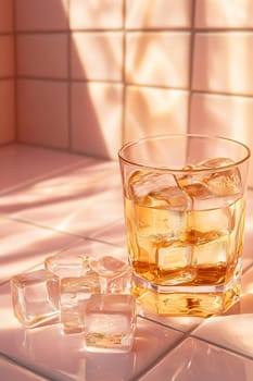 A glass filled with a golden beverage sits next to ice cubes on a light pink surface with warm sunlight casting soft shadows.