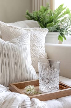 A stylish living room features a crystal glass on a wooden tray, surrounded by decorative pillows and indoor plants, creating a serene atmosphere.